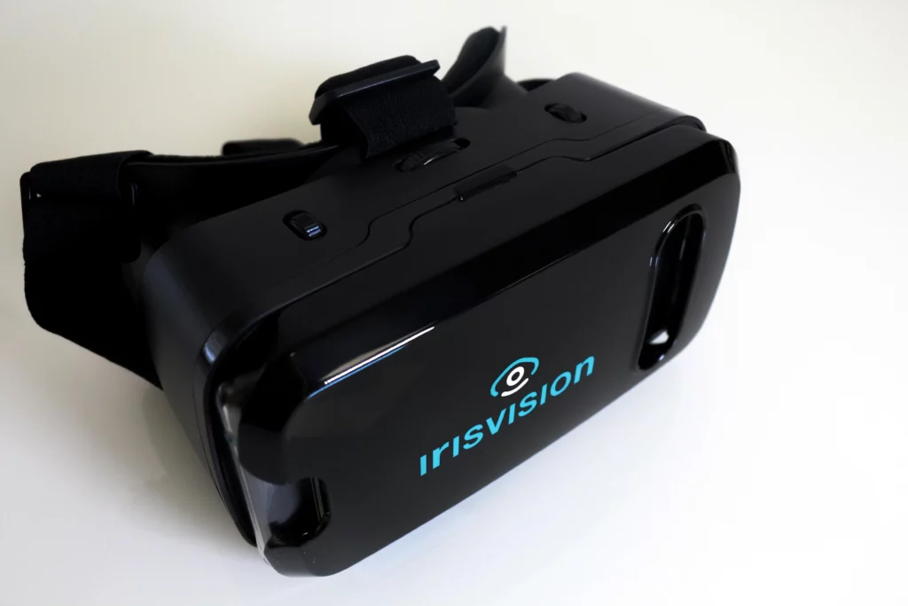 A black VR headset lies on a white surface, with adjustable head straps. The text "IRISVISION" is printed on the front in blue letters.