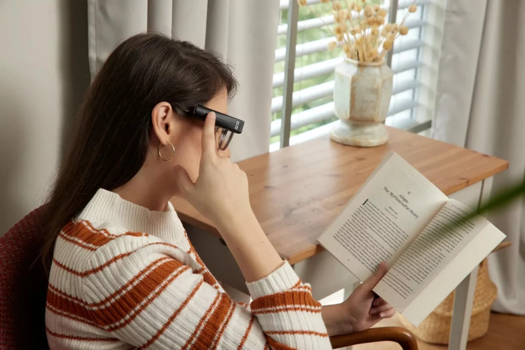 A woman wearing  glasses  with Orcam attached to one arm. She reads a book titled "The Mythical Story" near a wooden table with a vase of dried flowers, beside a window with blinds.